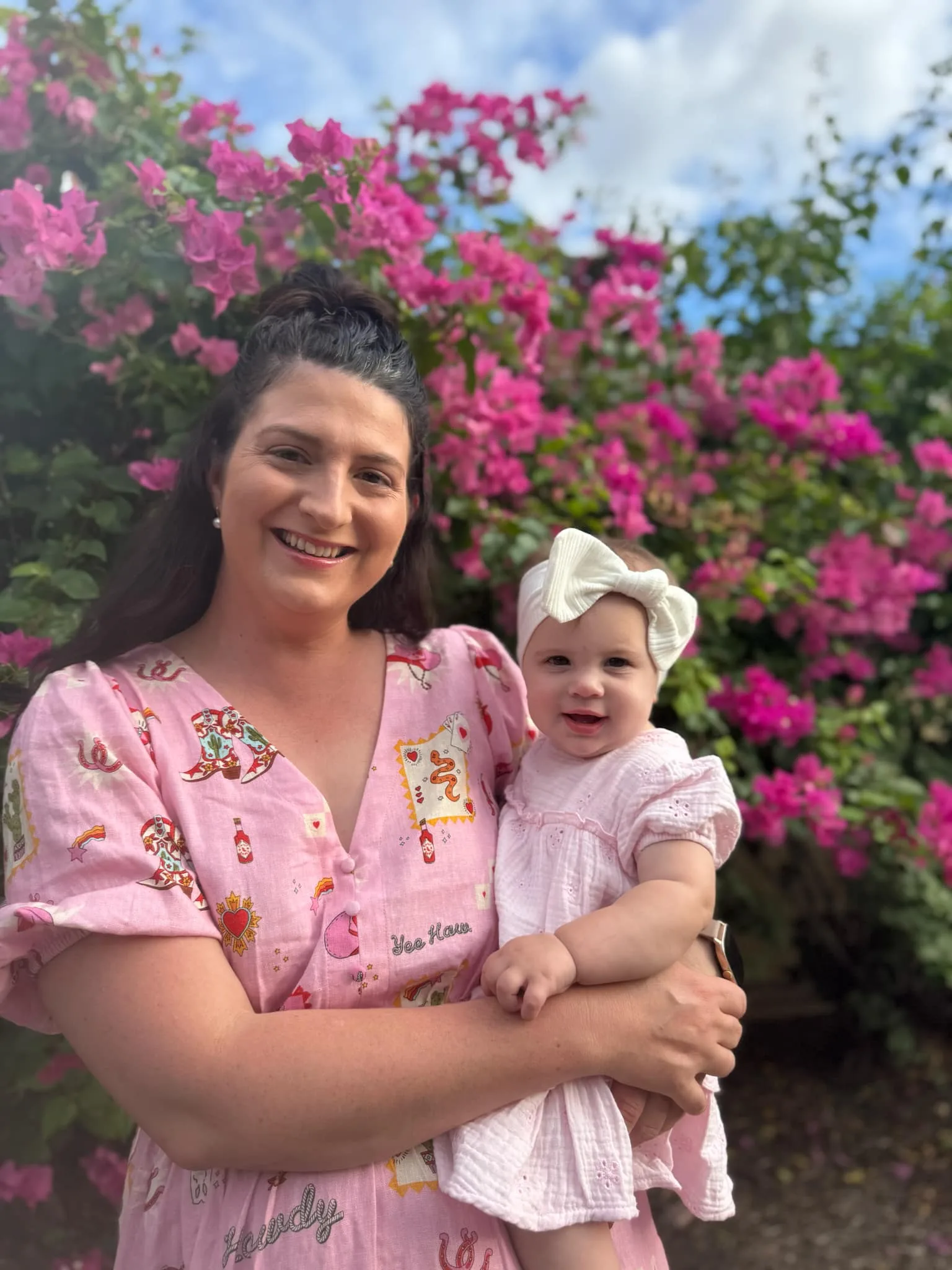 Woman in pink top holding baby with white bow in front of bush with pink flower.