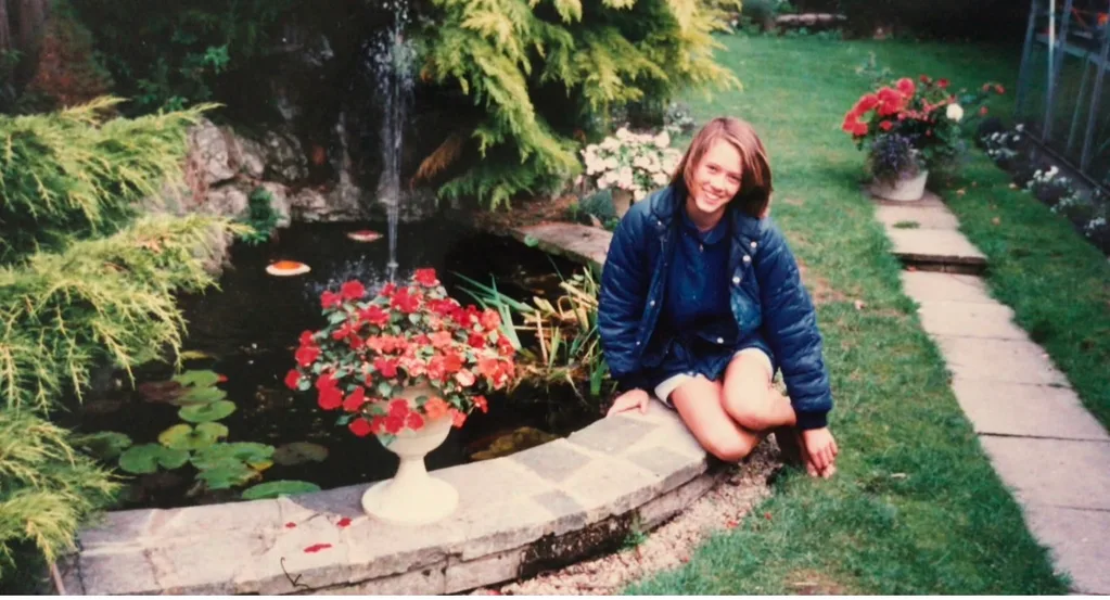 woman in blue jacket sat by a pond in a garden