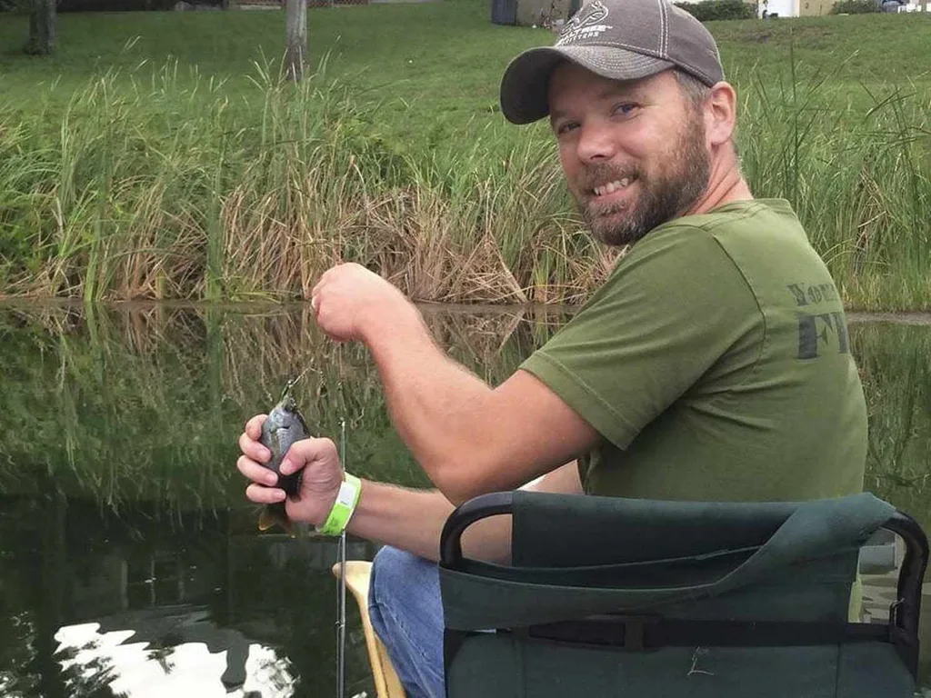 Michael Shaver smiling at the camera as he proudly holds a fish he's caught. He is wearing a grey cap, green shirt and blue jeans