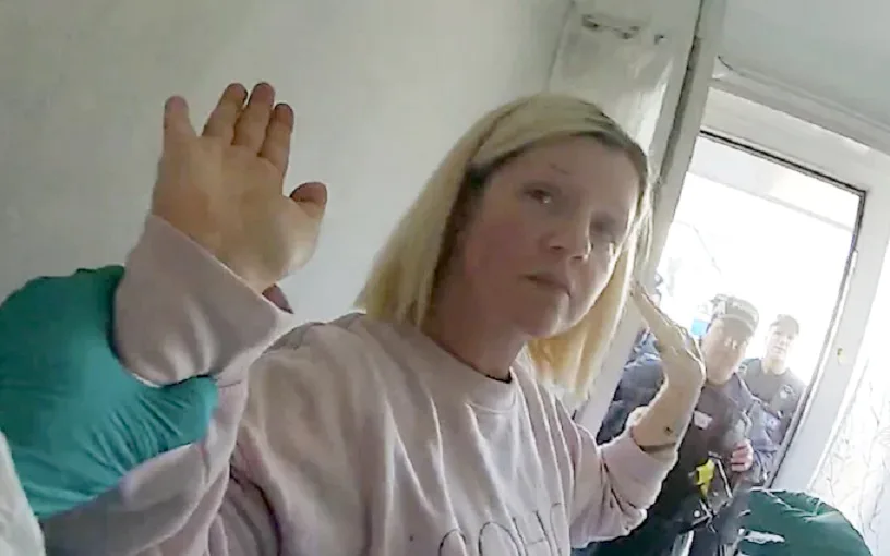 A blonde woman raises her hands in the air in surrender. The police can be seen standing behind her