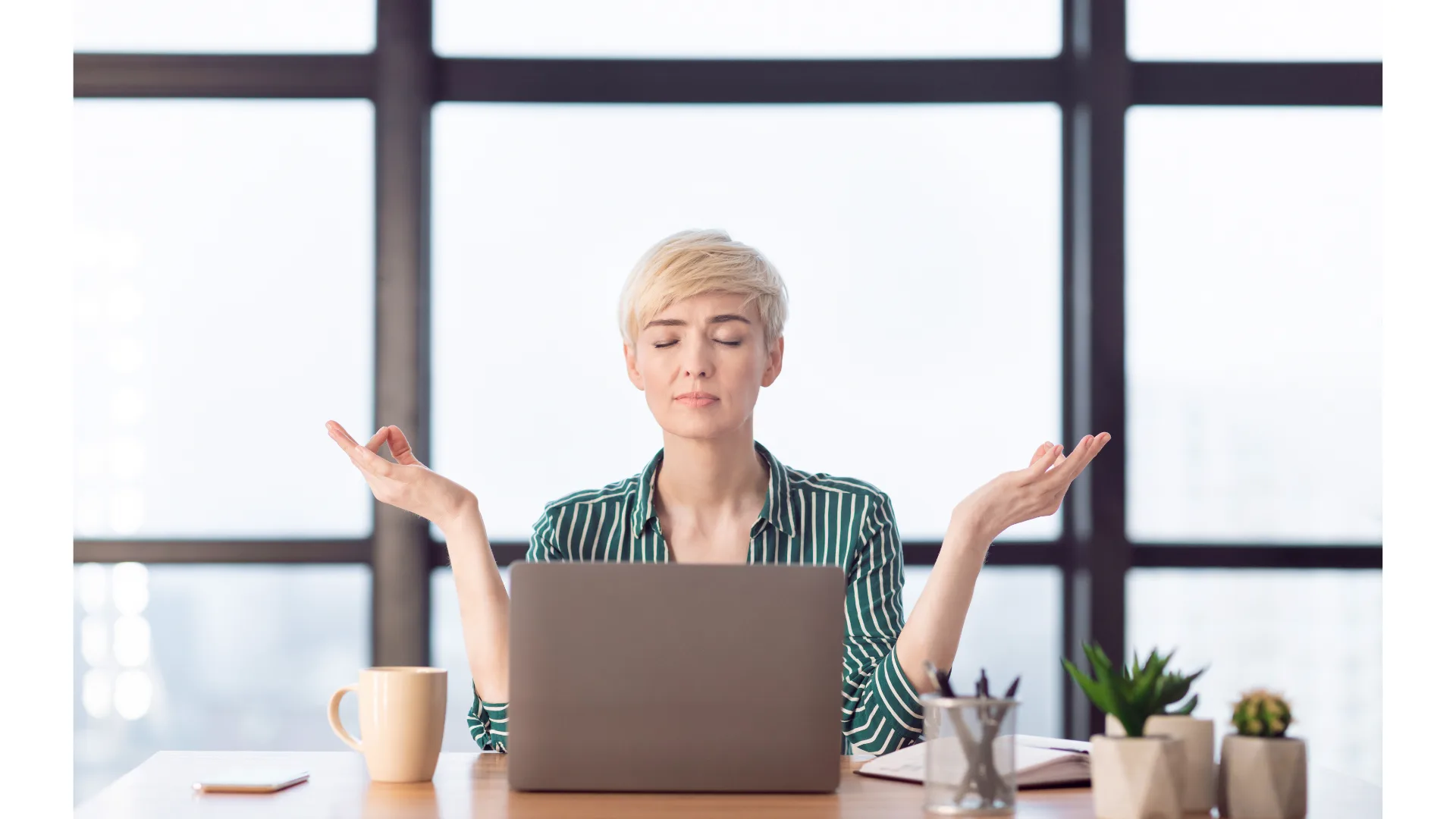 A short-haired woman seated behind her laptop closes her eyes as she meditates.