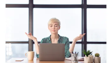 A short-haired woman seated behind her laptop closes her eyes as she meditates.
