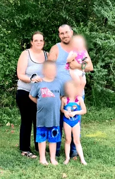 Image of family standing in front of green bush. She is wearing a striped black and white singlet and black pants, he is wearing a grey singlet. The three children have their faces blurred out for privacy