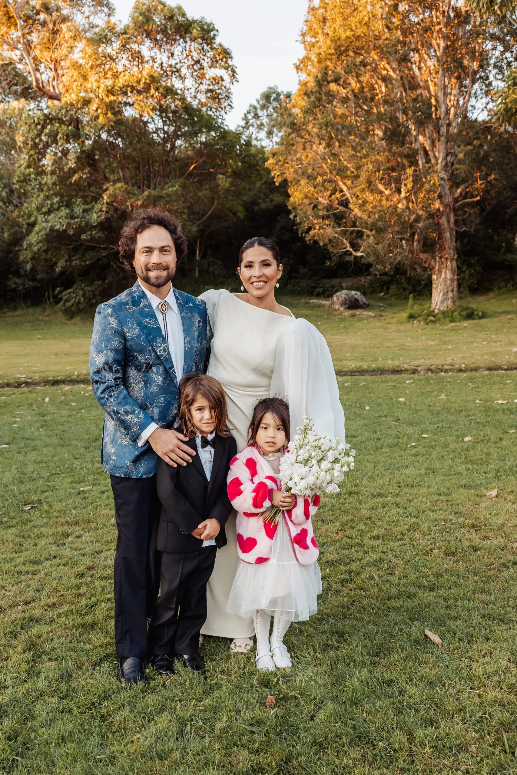 bride and groom with two children a boy and girl standing on grass. He is wearing a blue blazer. Her wedding dress has long billowing sleeves. Their son is wearing a black tuxedo and their daughter is wearing a white dress and a pink jacket with red hearts