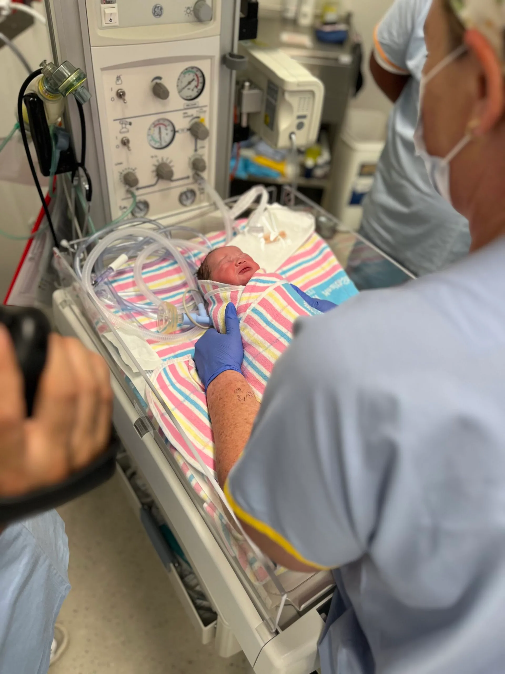 Tiny baby girl wrapped in pink, blue, yellow and white hospital balnket. A nurse is wearing glove and holding her.