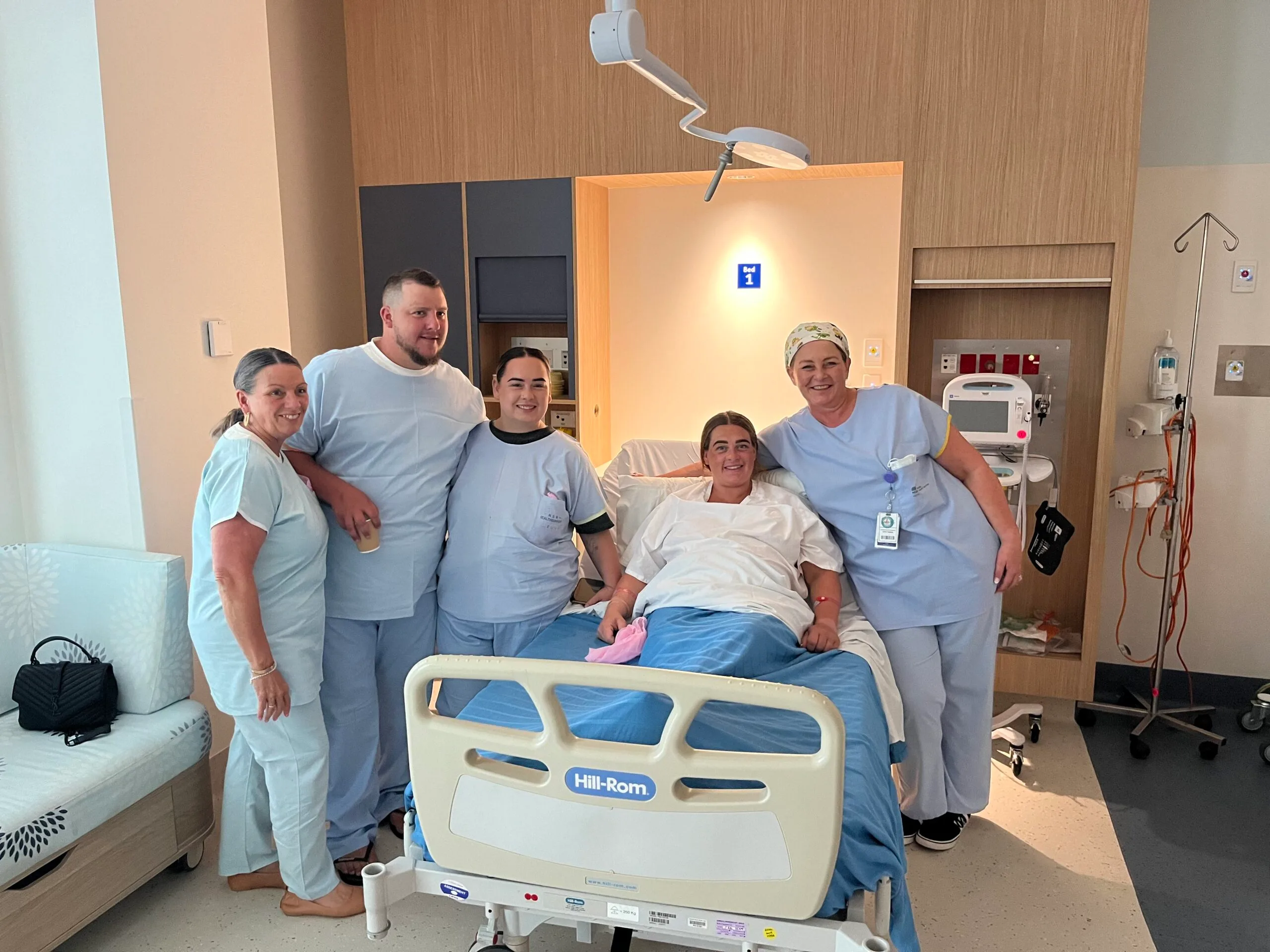 Group of people wearing blue scrubs around woman in hospital bed
