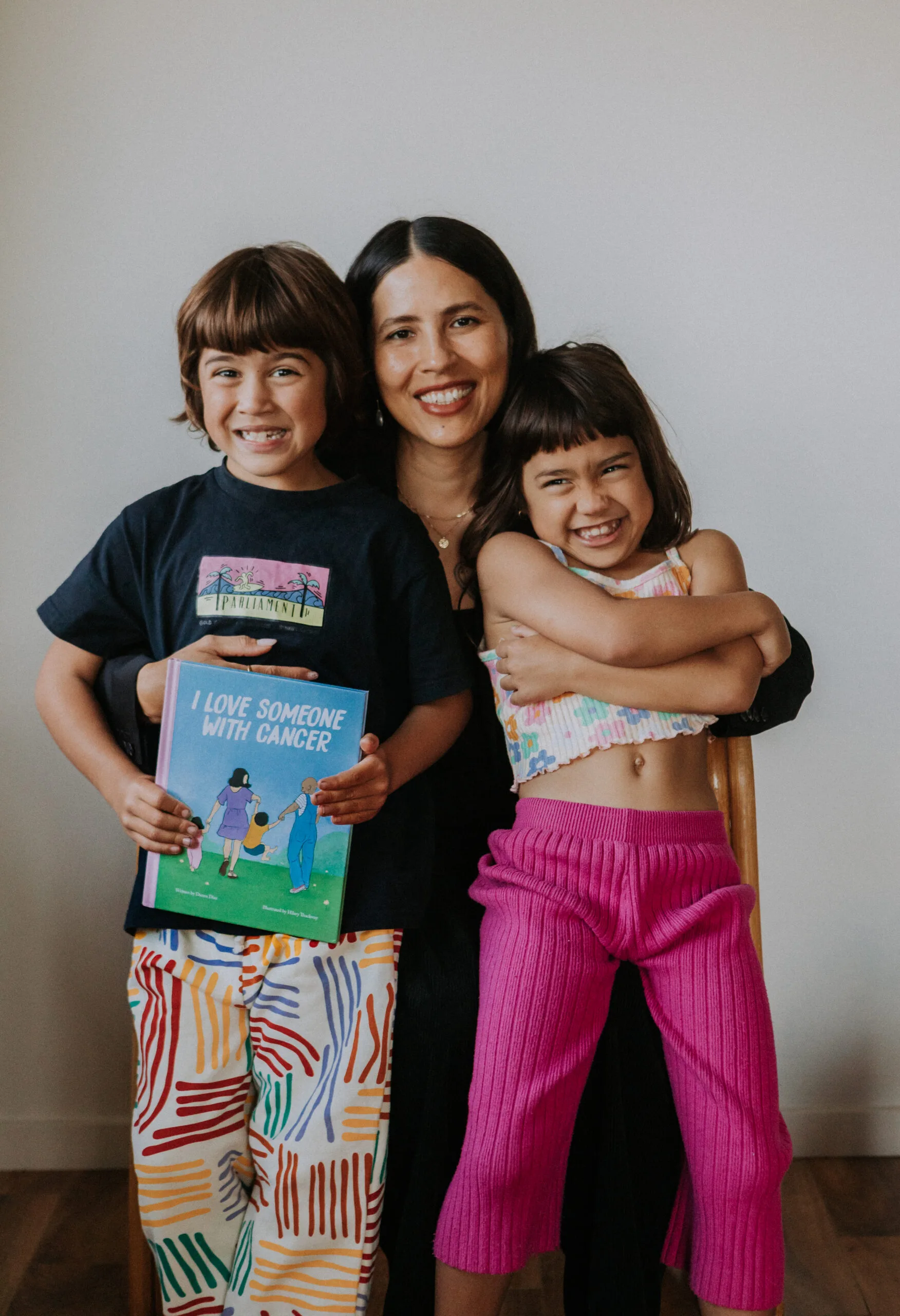 woman cuddling with two kids holding a book. She is wearing a black dress. Her son is wearing a black shirt and colourful pants. Here daughter is wearing a white floral top and pink pants