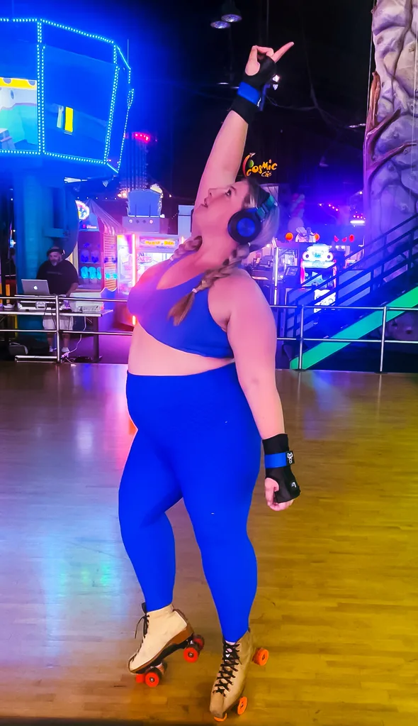 Connie when she started skating at her local roller skating rink. She is wearing a bright blue crop top and tights, as well as black wrist guards, headphones and white roller skates