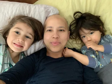 Mum in black top smiling for the camera with her two little children by her side. She has no hair