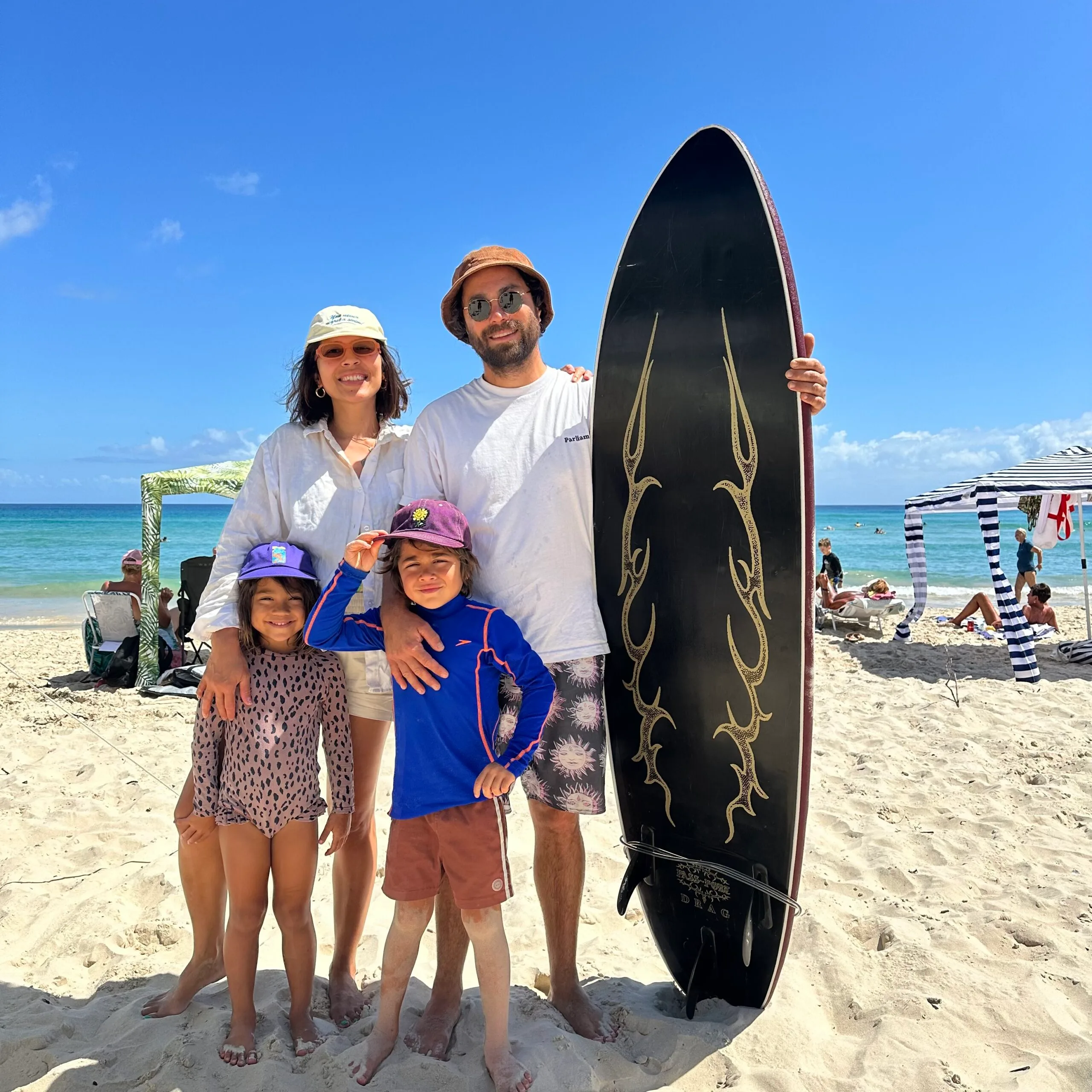 Woman husband and two children at the beach. She is wearing a white long sleeve shirt with cream shorts and a yellow hat. He is wearing a white shirt and black shirts with suns on them. He is holding a black surfboard. The kids are wearing colourful swimwear and purple hats.