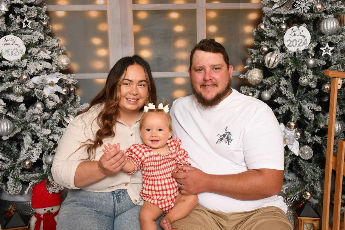 Couple sit with their baby in front of christmas trees, woman is wearing beige shirt and man ins wearing white tshirt, baby is dressed in red checked outfit