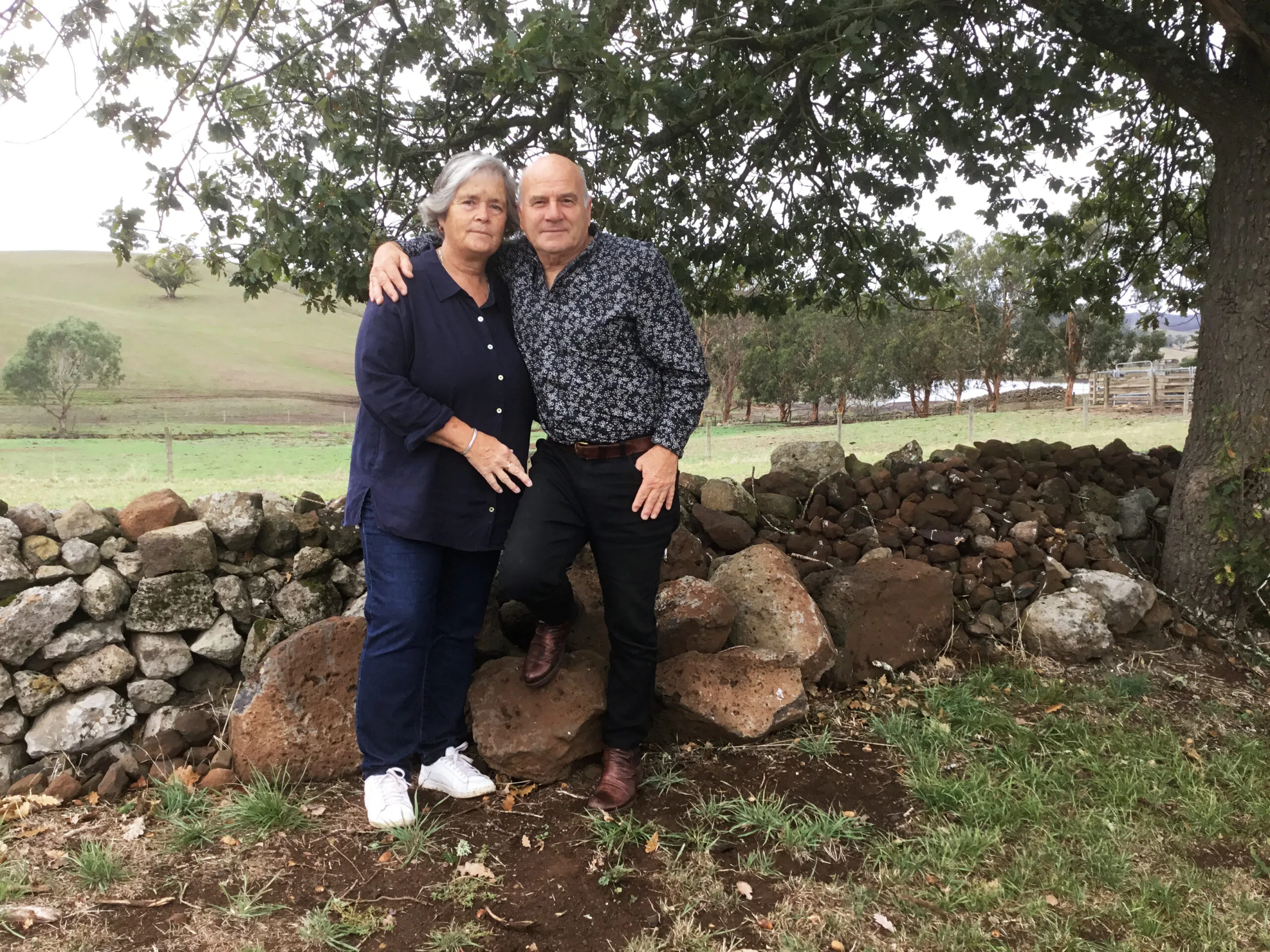 Couple sitting on a rock. He is wearing a grey business shirt, black pants and brown boots. She is wearing a navy blue long-sleeve shirt, dark blue jeans and white trianers