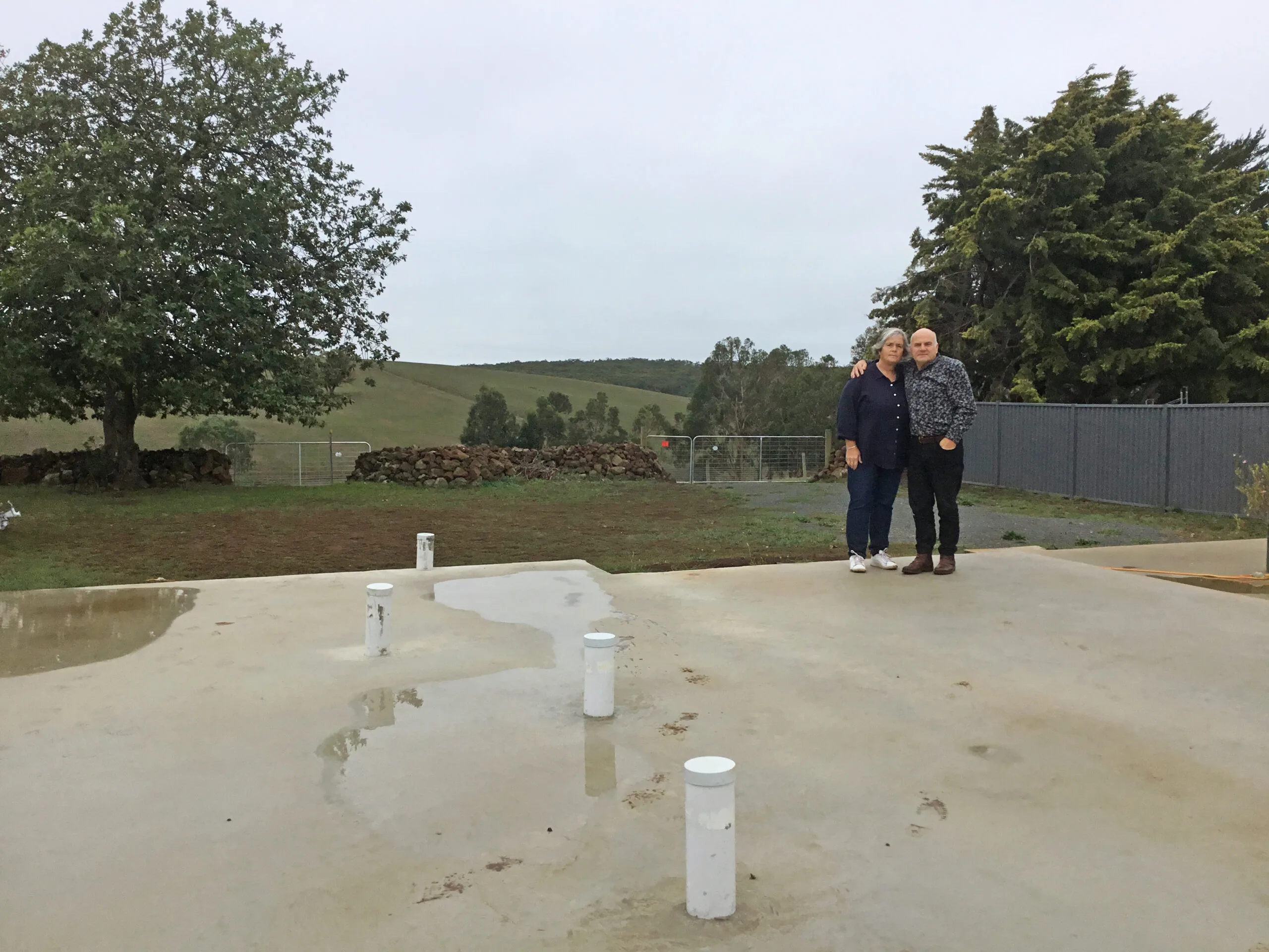A man and woman standing on concreate slab. He is wearing a grey business shirt, black pants and brown boots. She is wearing a navy blue long-sleeve shirt, dark blue jeans and white trianers