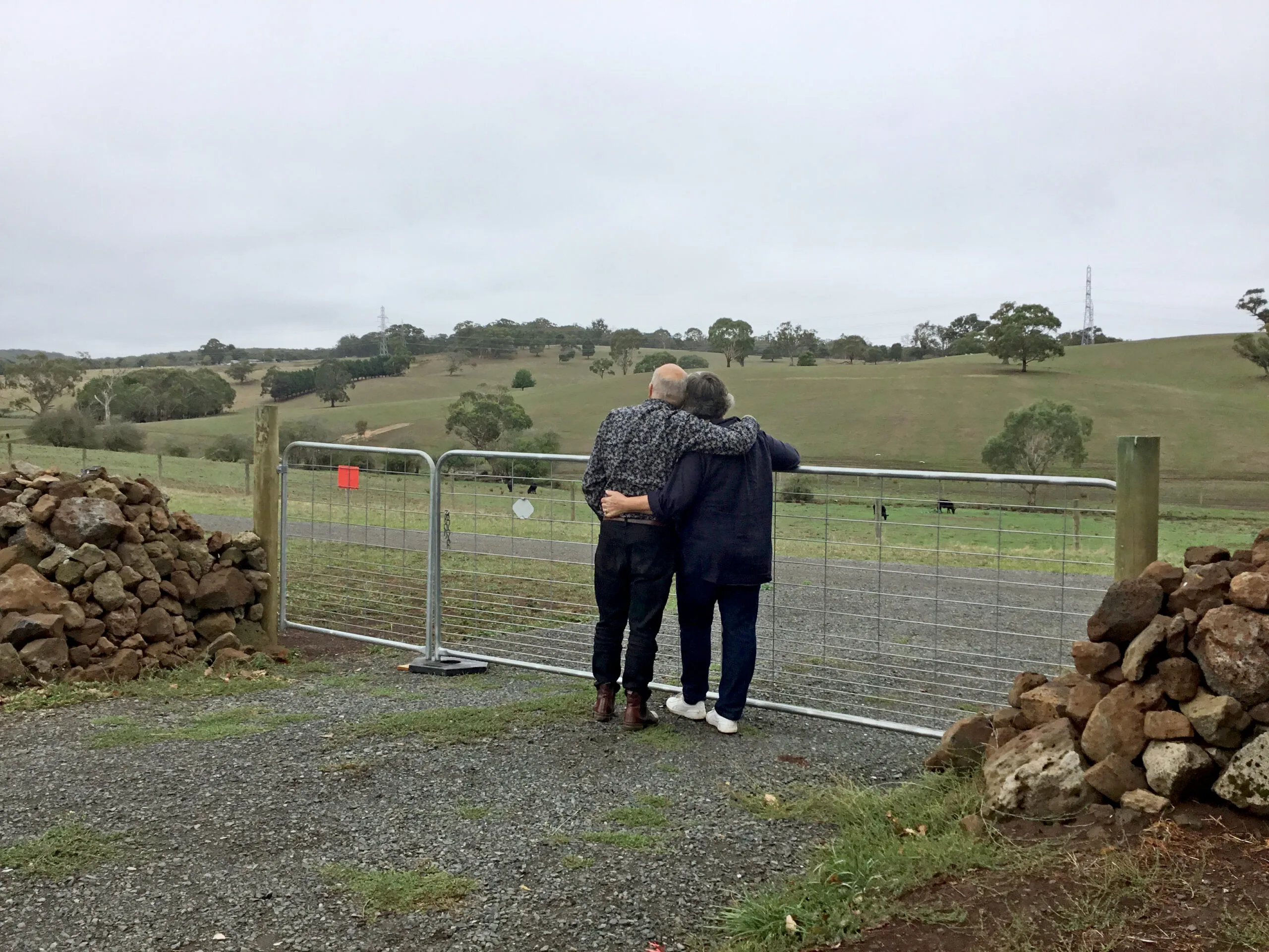 Couple arm in arm, backs to camera looking out over rolling hills. He is wearing a grey business shirt, black pants and brown boots. She is wearing a navy blue long-sleeve shirt, dark blue jeans and white trianers