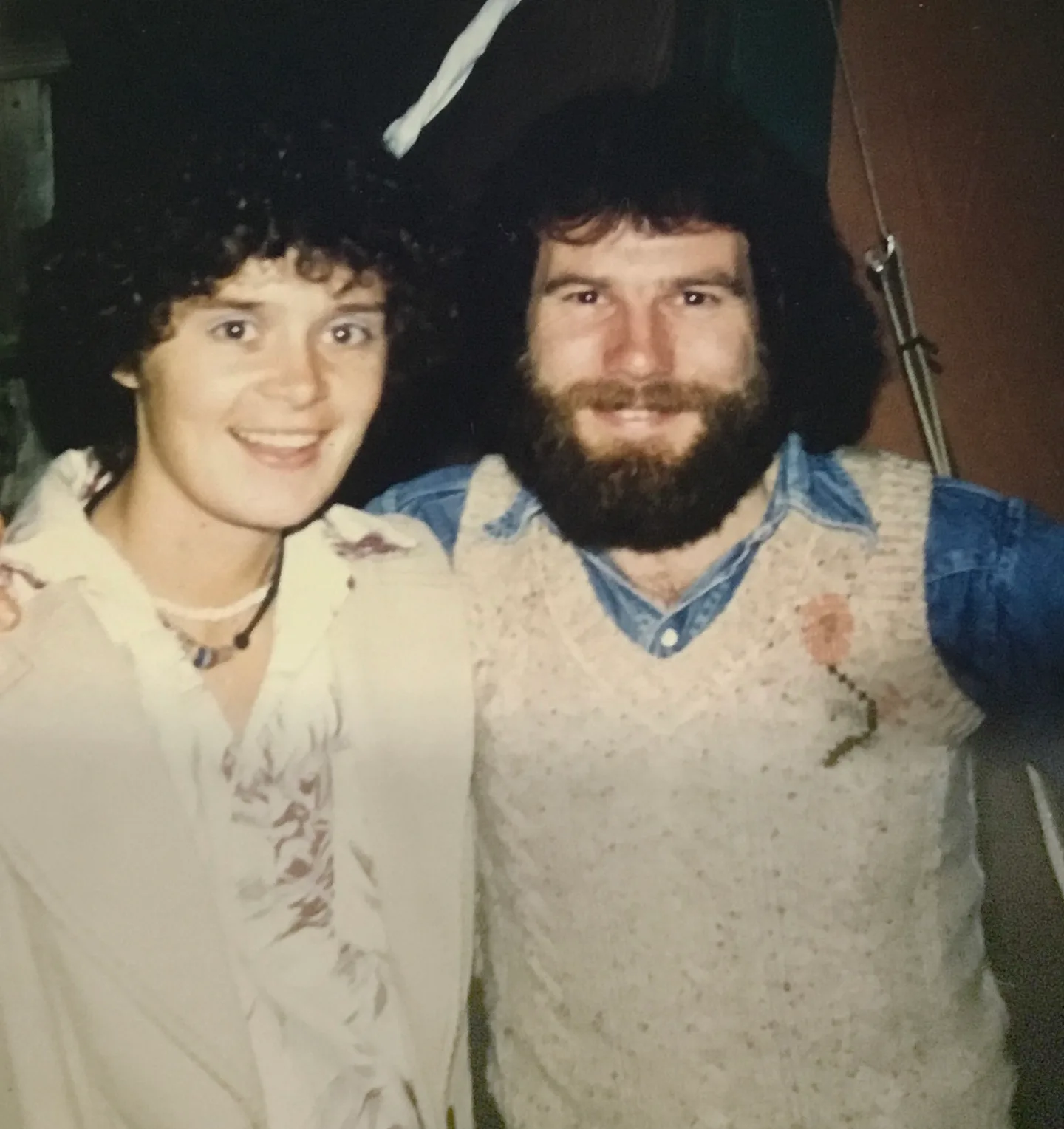 Retro photo of couple smiling. She has short brown curly hair and is wearing a white long-sleeve blouse. He has long brown hair and a brown beard. He is wearing a denim shirt with a beige wool vest over the top. He has his arm around her
