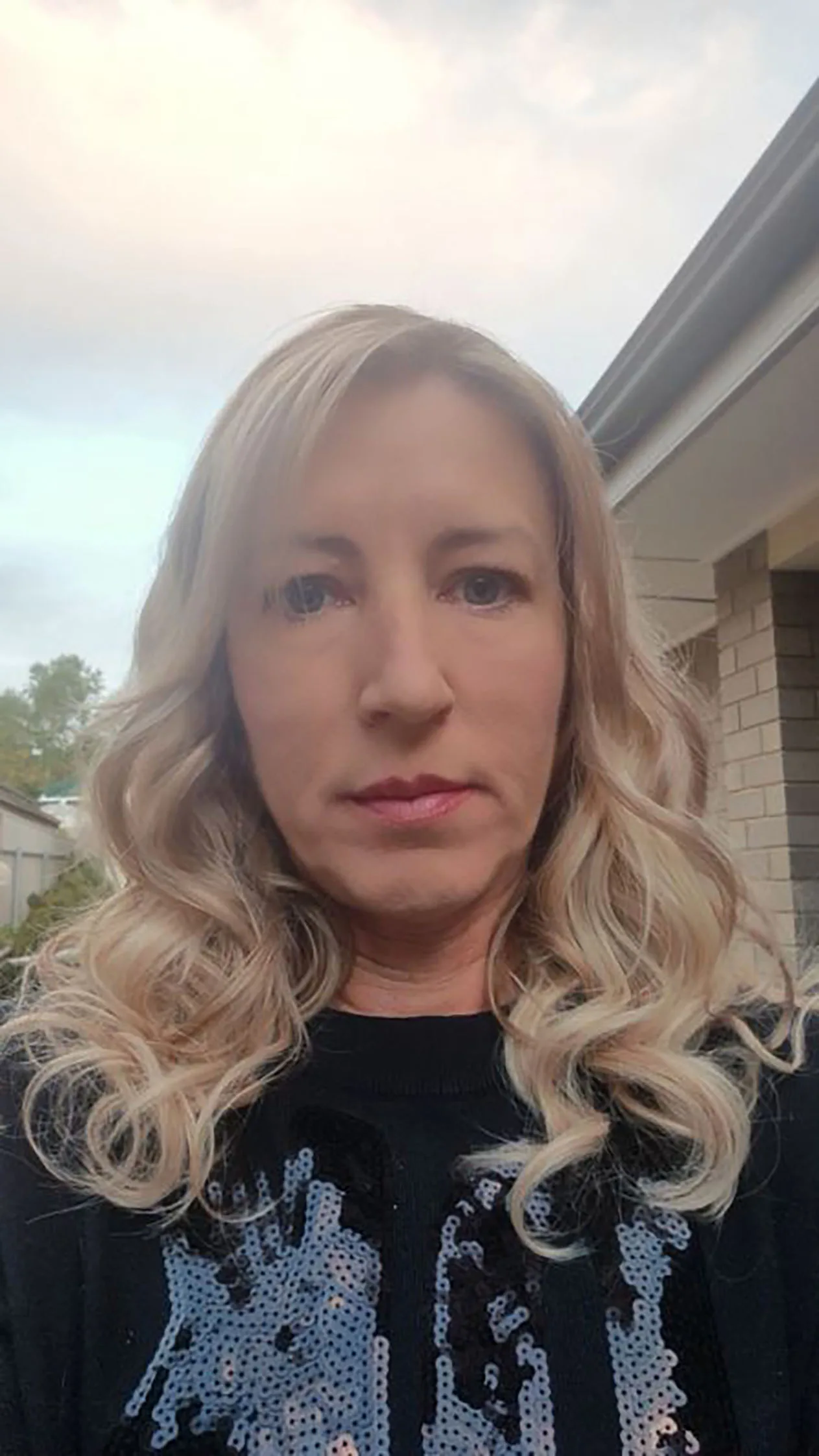 A recent picture of abuse survivor Kylie Bailey outside of a house. She has long blonde curly hair and is wearing a black top with silver sequins
