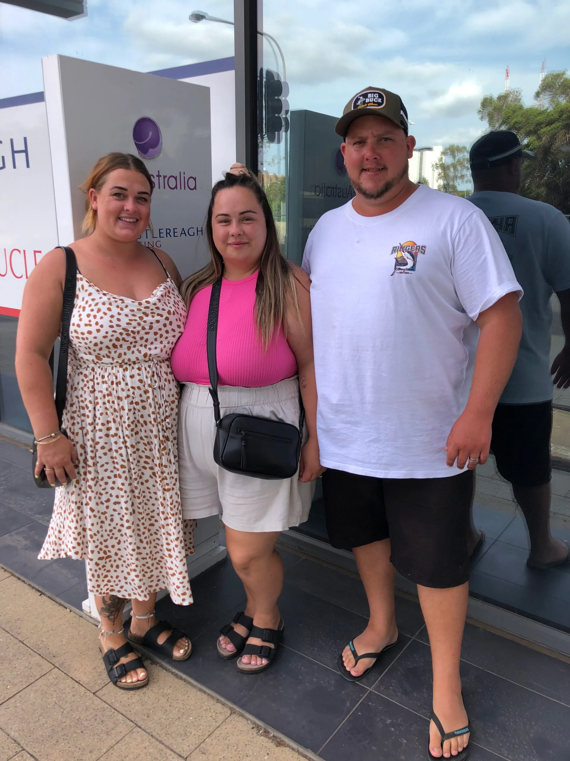 woman in pink top standing with her sister in white and brown spotted dress and her husband in white t-shirt and cap