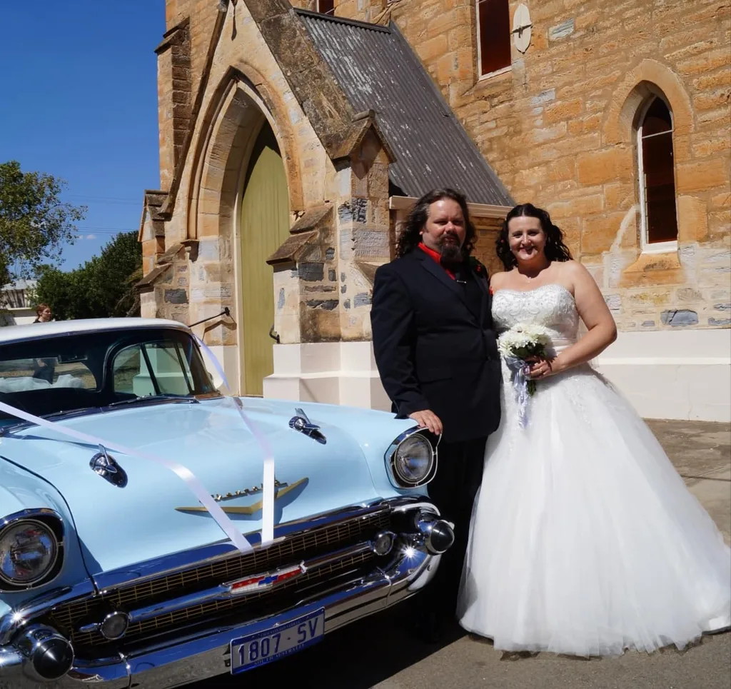 Man and woman on their wedding day standng in front of a vintage baby blue car. She is wearing a strapless lace wedding dress and he is wearing a black suit with a red shirt