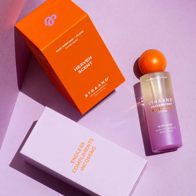 Colourful purple and orange beauty product packaging against a light purple background. Product is hair perfume