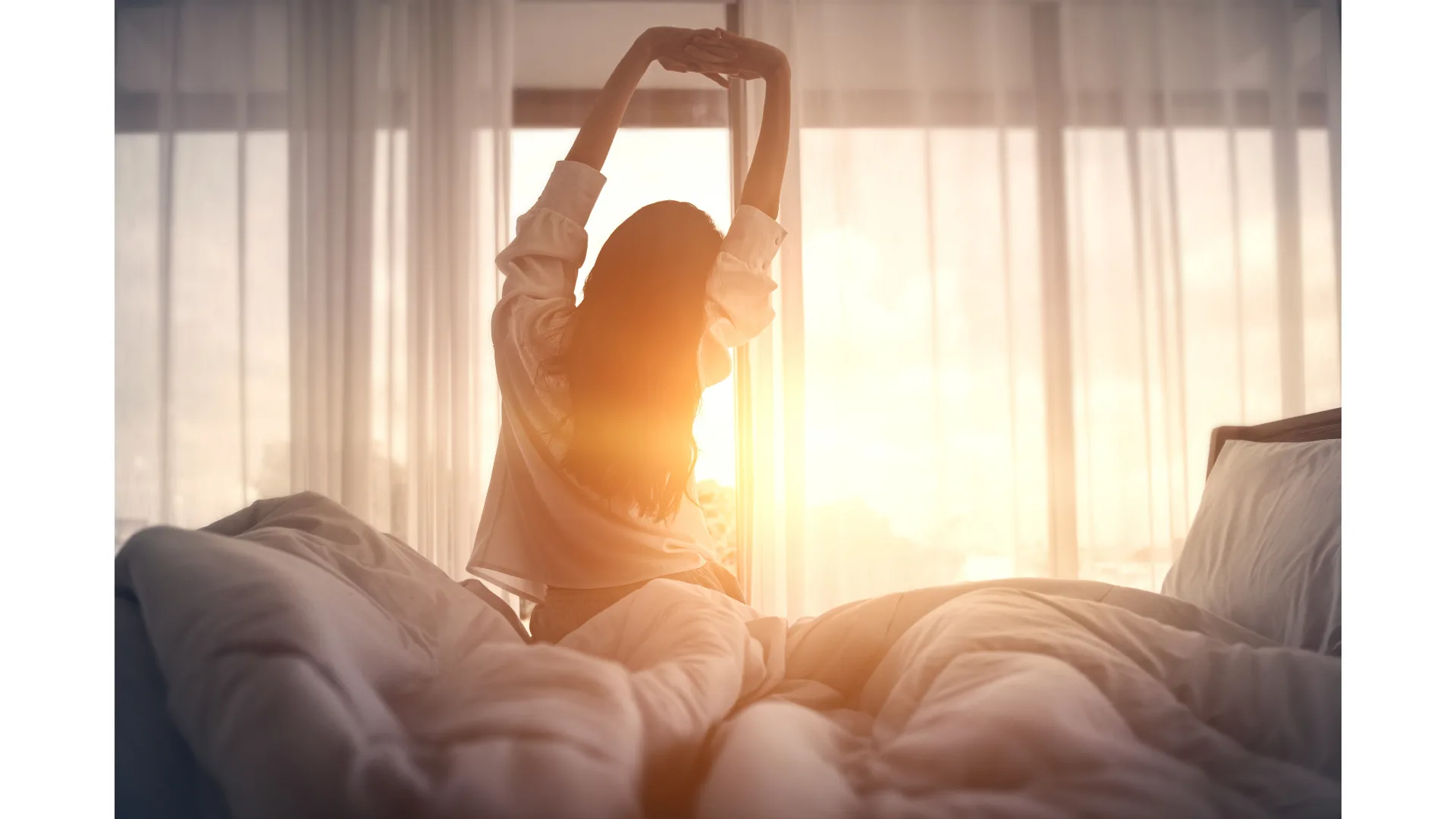 A woman sits on the edge of her bed and stretches as the sun rises outside her window.