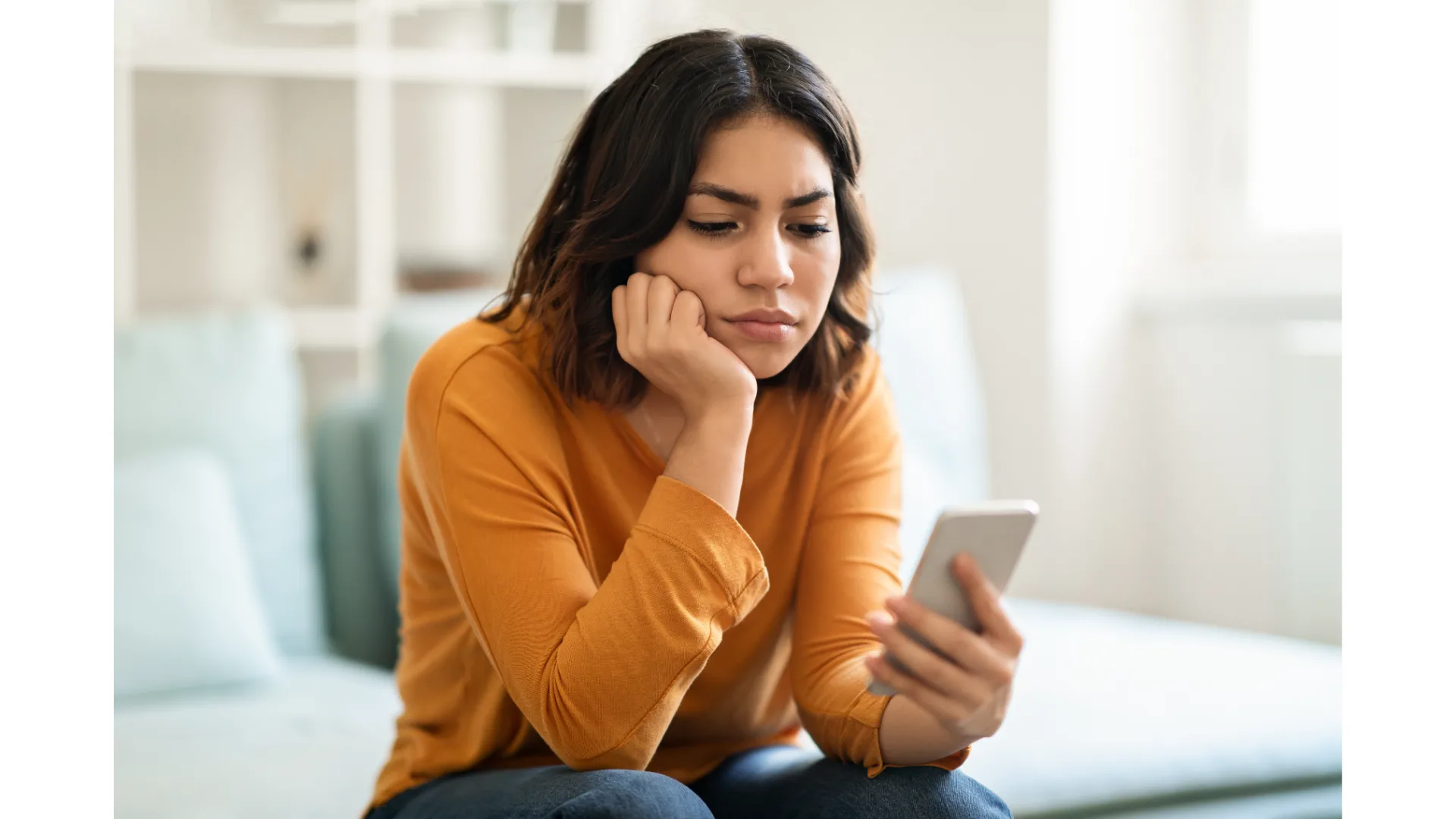 A brunette woman looks frustrated while sitting on a bed and staring at her phone.