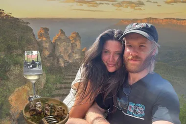 A couple (man and woman) against a backdrop of the Blue Mountains (two visuals overlapping - one of the Three Sisters and the other is a walking track). Next to them on the left is a glass of white wine and a plate of halloumi.