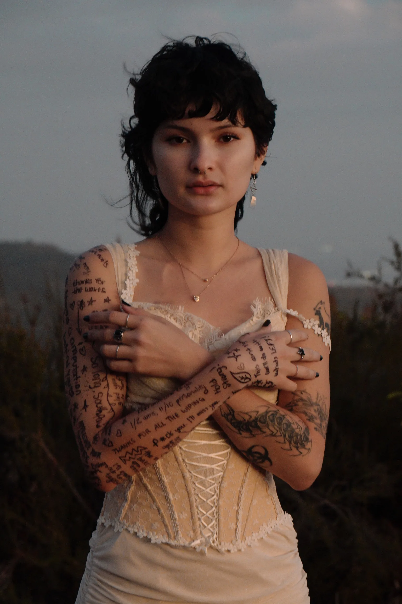 Young woman with short black curly hair in white lace top. She has tattoos on her left arm and her right arm is covered in farwell messages drawn on by her friends