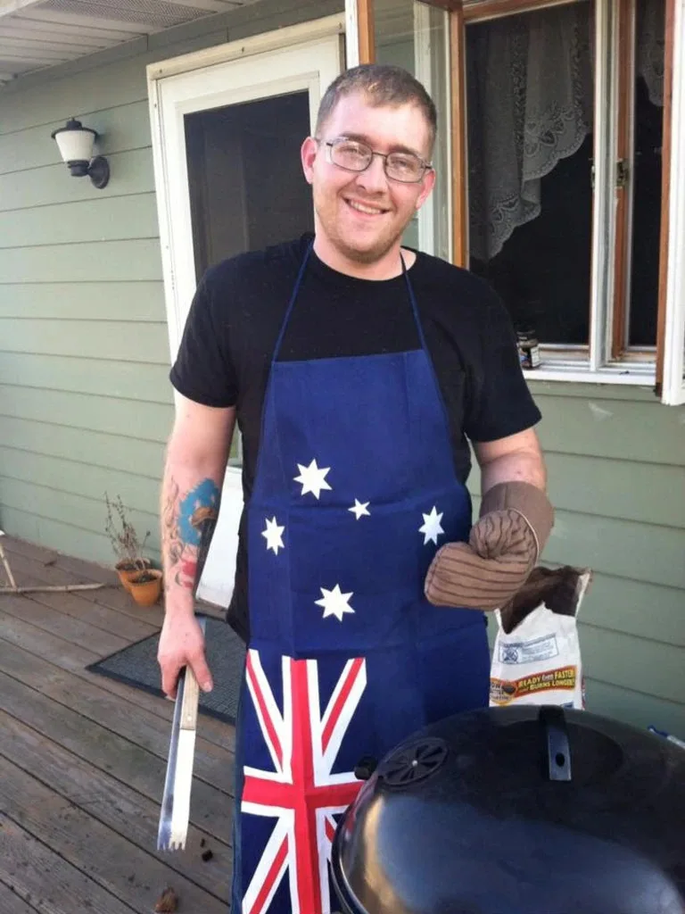 A man is standing next to a webber barbecue. Is wearing glases, a black shirt and an Apron with the Australian flag