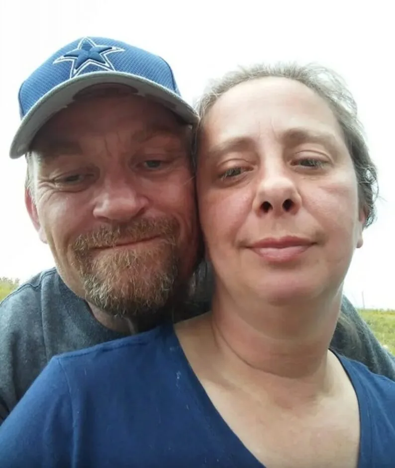 Man and woman smiling in a field. He is standing behind her hugging her from behind wearing a navy cap, she is smiling