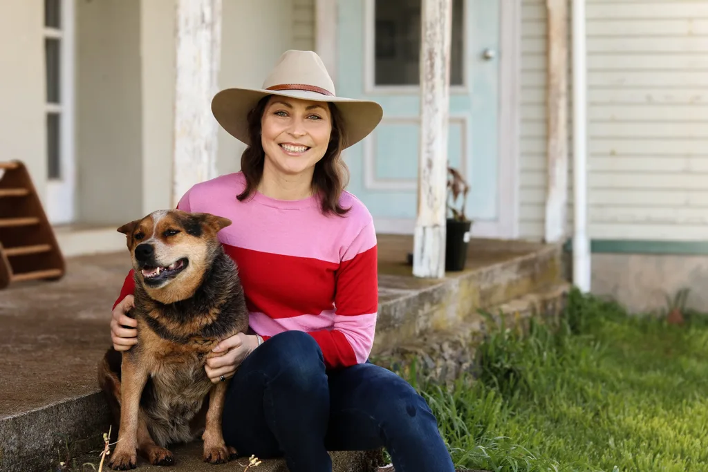 A woman smiling with a dog. She is wearing a hat and a pink and red shirt. The background is a cartoon style picture of the countryside.