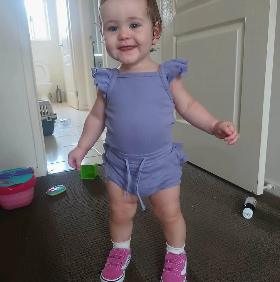 Toddler smiling for the camera. She is wearing a lilac top and shorts and pink shoes