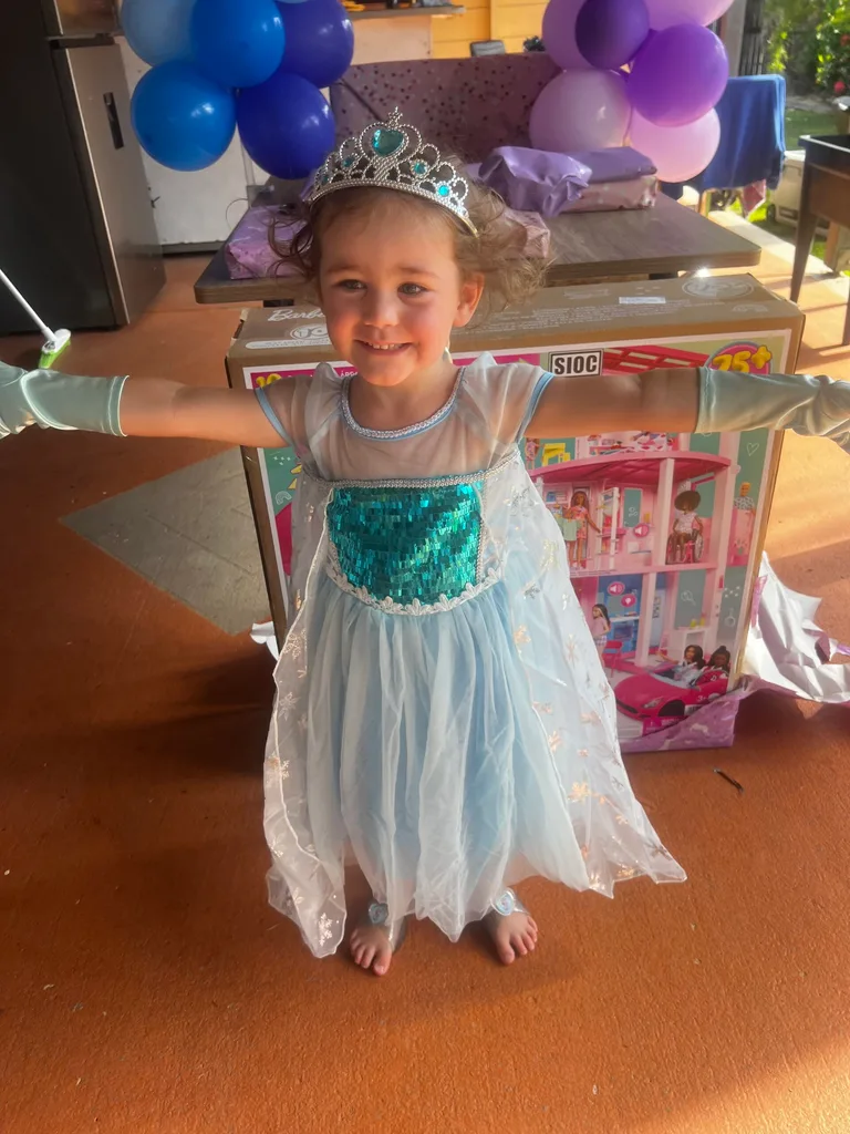 A toddler in a green and blue princess dress. She is wearing a silver and green cross and matching blue gloves