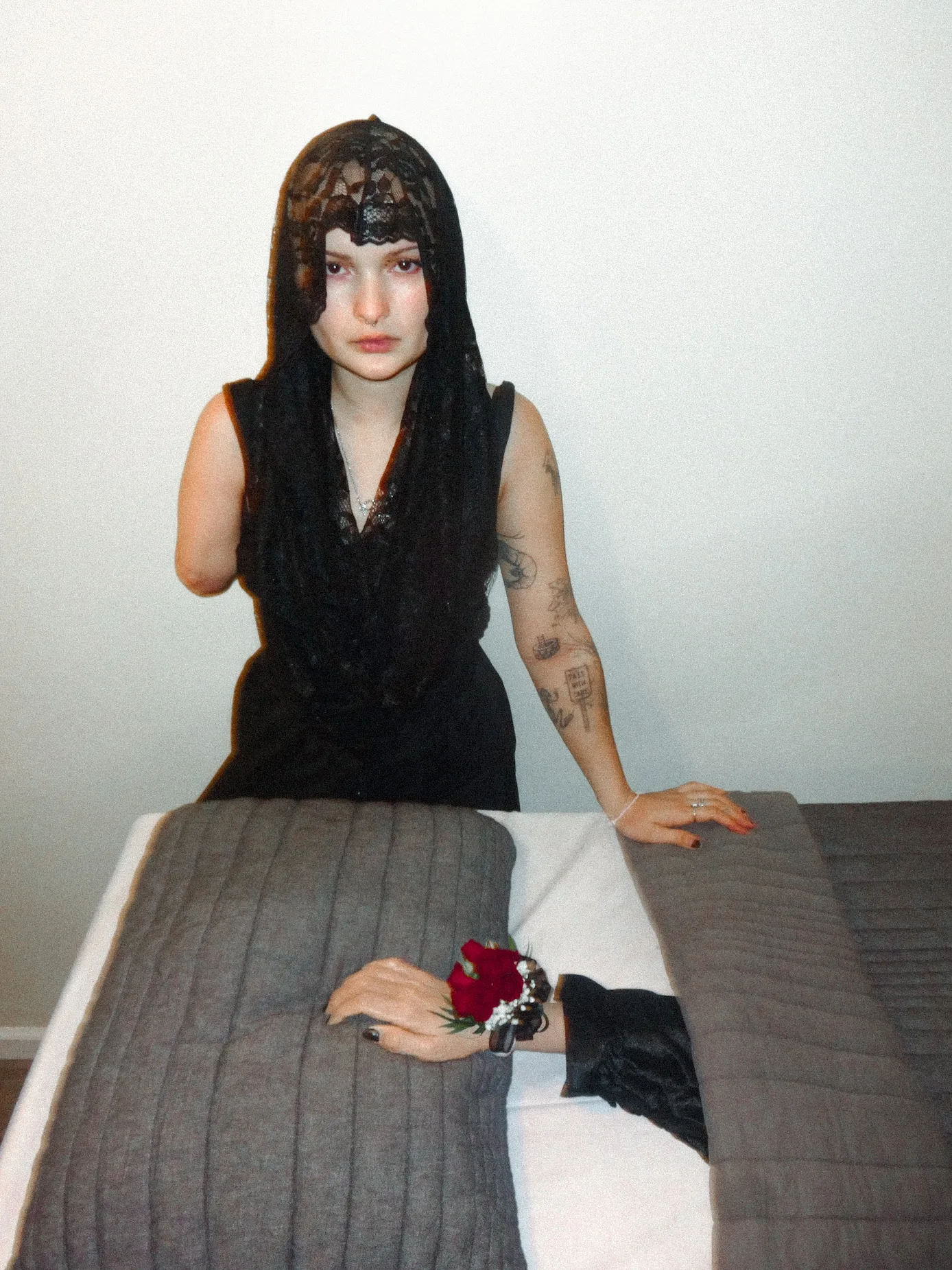Young girl in black lace top with attached hood. She is leaning on a bed with her amputated arm tucked under a blanket. The nails are painted black and it is wearing a red corsage