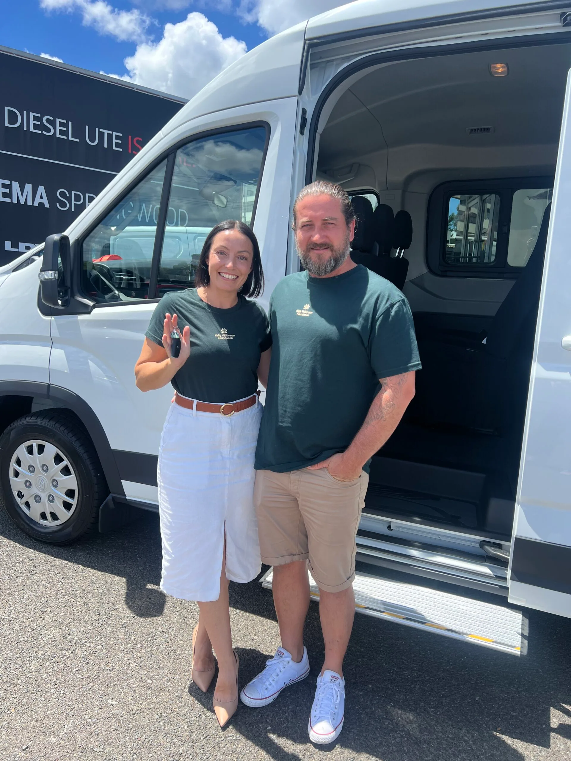 A couple standing in front of a white van. The woman on the left is wearing a dark green t-shirt, white midi skrit and brown belt. The man on the right is wearing a matching green t-shirt and beige shorts