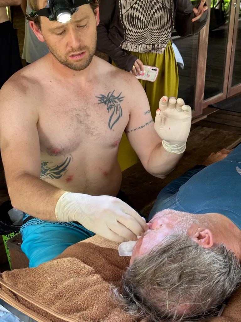 shirtless man with headtorch on in blue shorts and white gloves stitching face of man with grey hair lying odwn.
