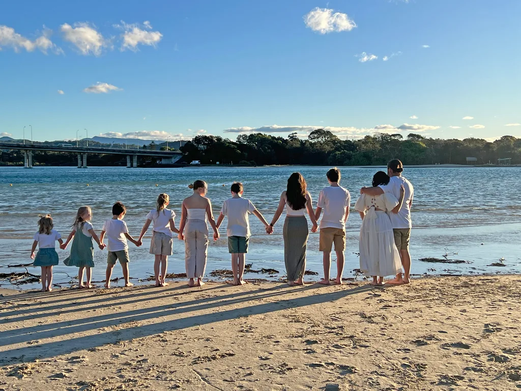 Two parents standing in a warm embrace on a beach. Eight children stand next to them hand-in-hand. They are all wearing white t-shrits and have their backs to the camera