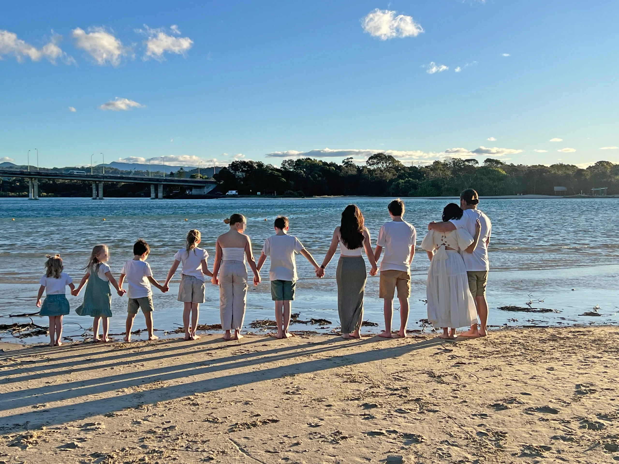 Two parents standing in a warm embrace on a beach. Eight children stand next to them hand-in-hand. They are all wearing white t-shrits and have their backs to the camera