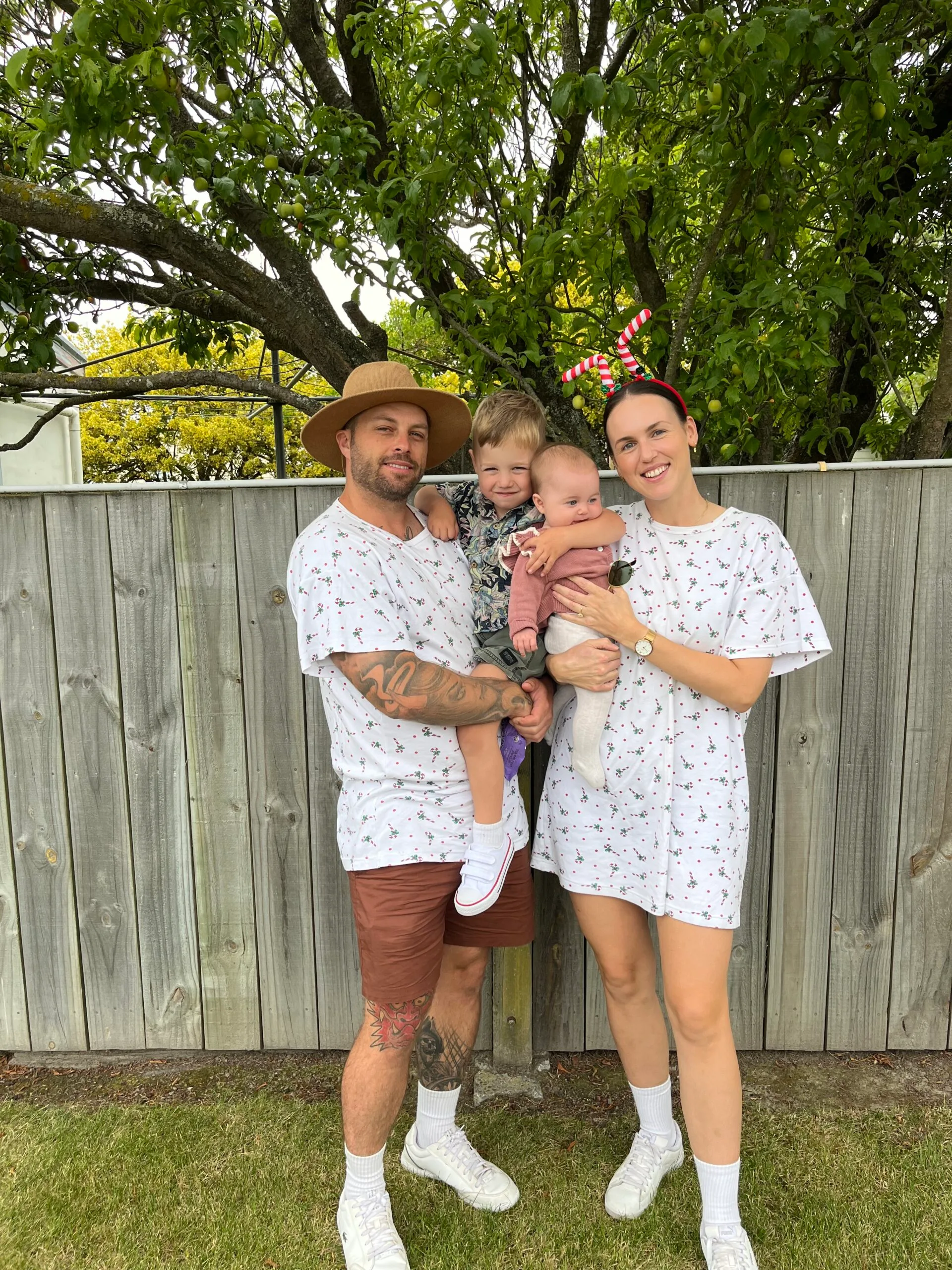 Family of four standing in backyard against a fence. The dather on the left is holding a son, the mother is holding a baby girl and they are wearing matching christmas outifts.