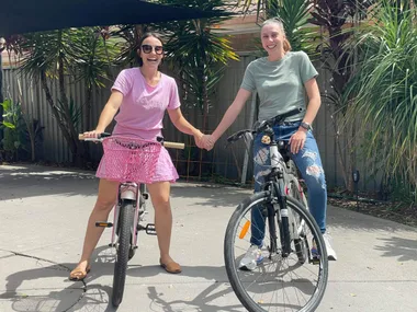 Sisters sitting on bikes and holdng hands. The woman o the right is wearing a pink shirt and skirt. The woman on the left is wearing a sage green shirt and blue jeans