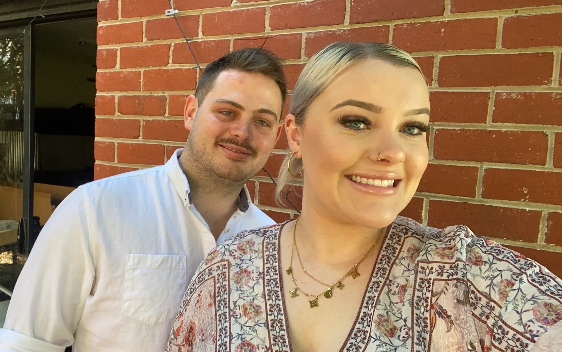 woman wearing pailsey dress smiling a the camera with her boyfriend he is wearing a white shirt. There is a brick wall behind them