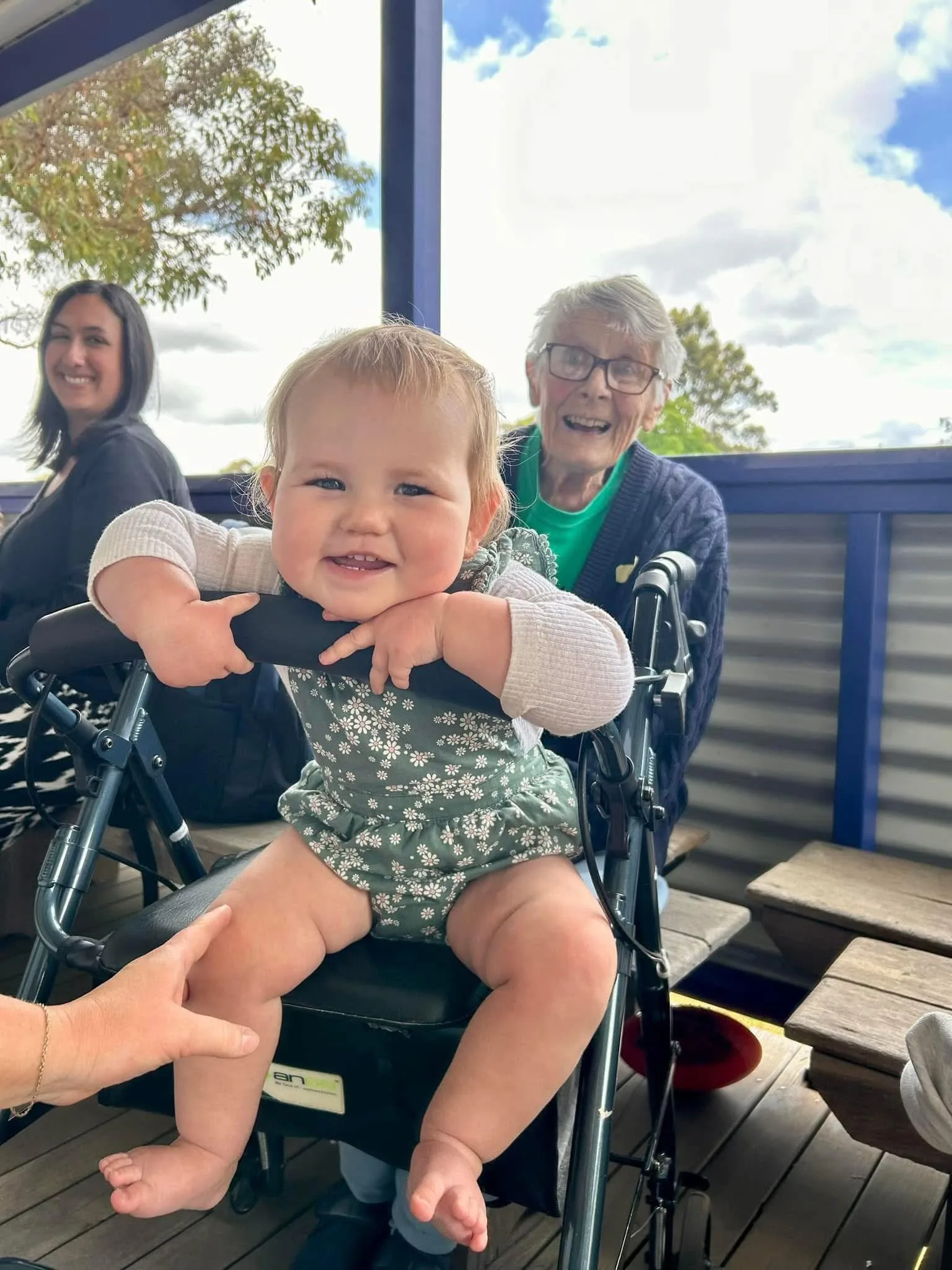 Image of baby wearing floral blue onesie and sitting on nanas walker with nana wearing green top