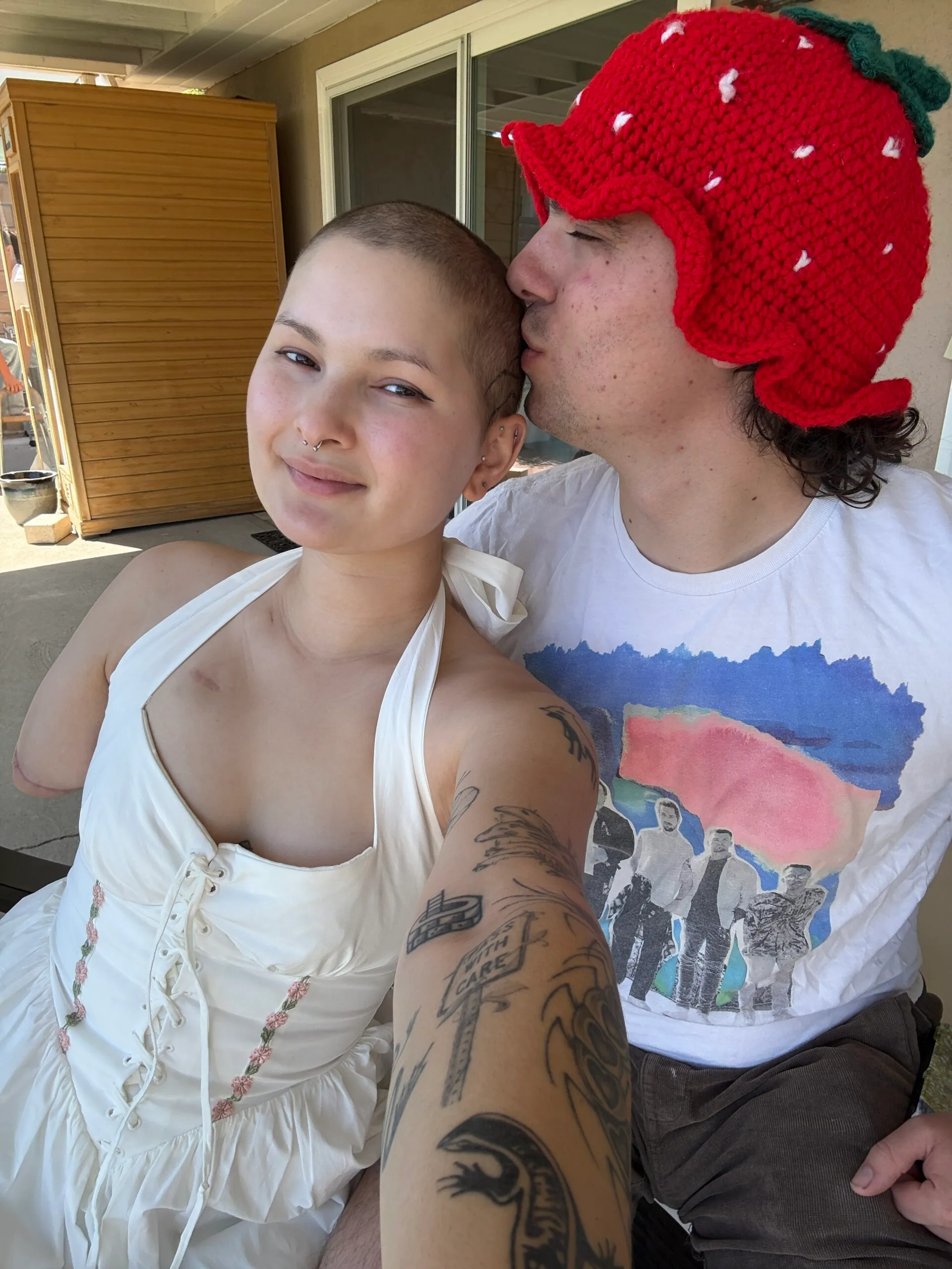 Young girl wearing a white halterneck top smiles for the camera. Her boyfriend is kissing her head. He is wearing a red knitted beanie