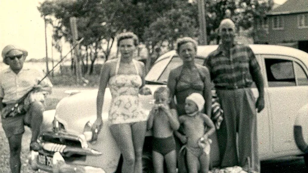 And old black and white photograph of some adults and children, They are outside and standing in front of a car. Their fashion is retro. They are all standing as a group and looking towards to camera.