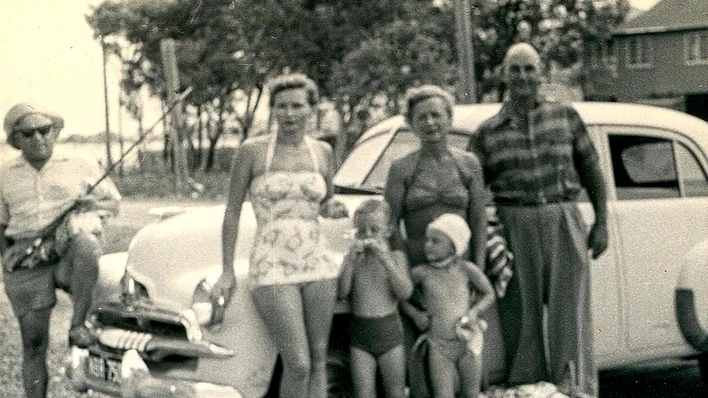 And old black and white photograph of some adults and children, They are outside and standing in front of a car. Their fashion is retro. They are all standing as a group and looking towards to camera.