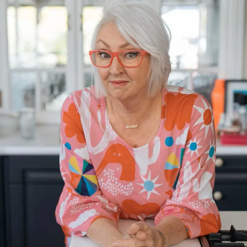 A modern photograph of a woman in her senior years. She is looking at the camera with a friendly expression. She is wearing red glasses and a colourful top with shades of blue, white, orange and pink.