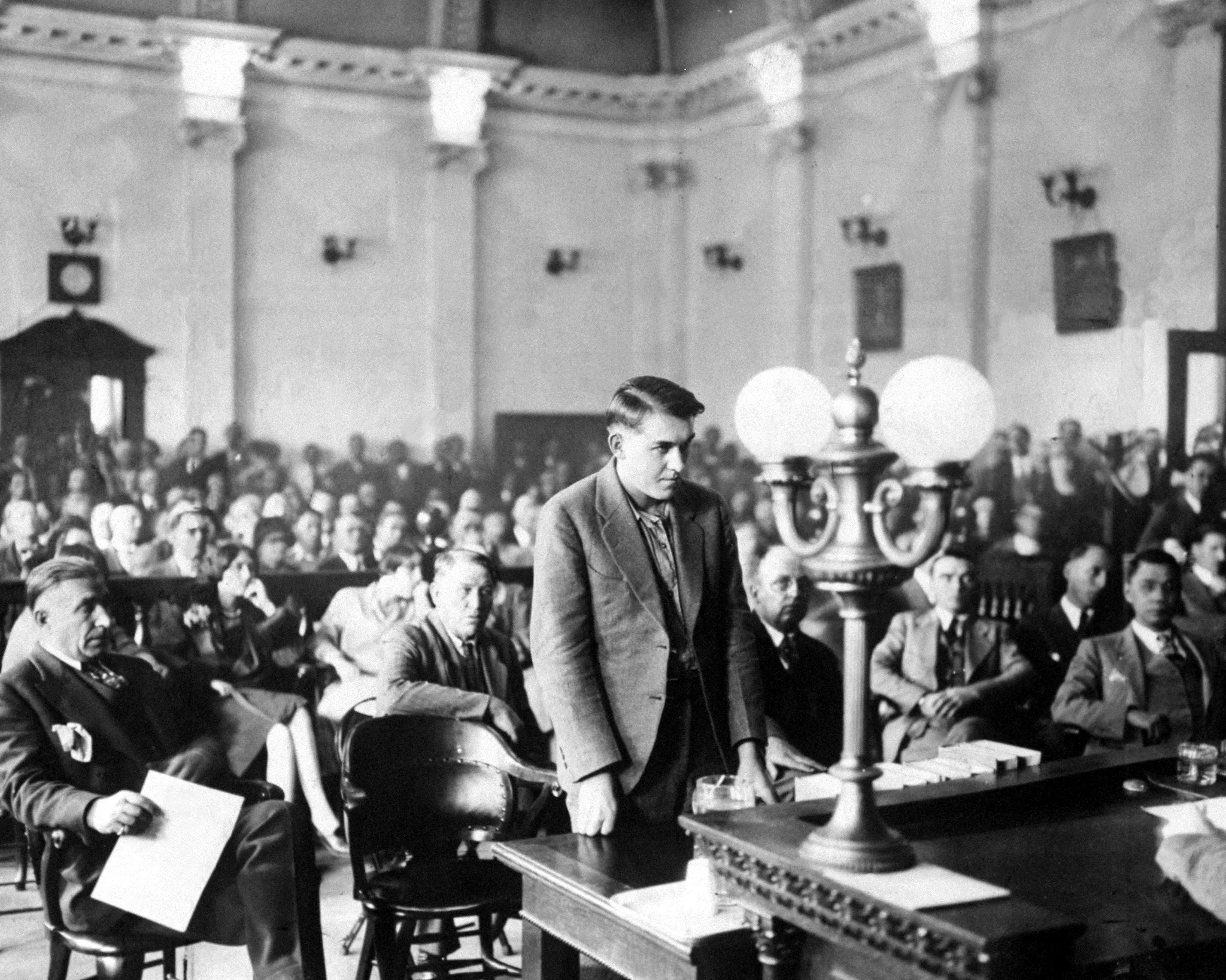 Man stands in front of crowded gallery in a 1920s courtroom 