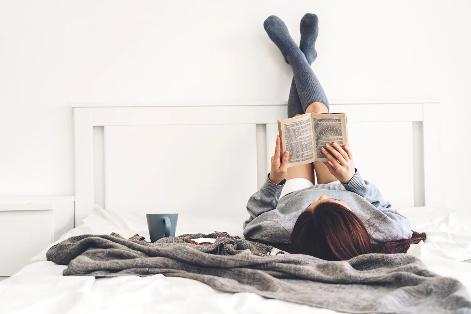 Young woman on bed with her legs up against the wall, reading a book with a cup of tea and biscuits