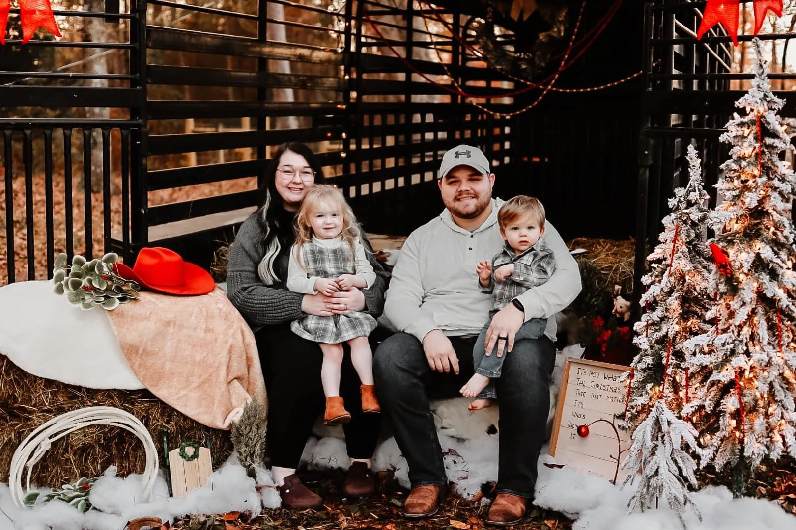 A family sitting for a Christmas photo. There is a tree and festive lights, the little girl sits on her mothers lap and young boy on the fathers 