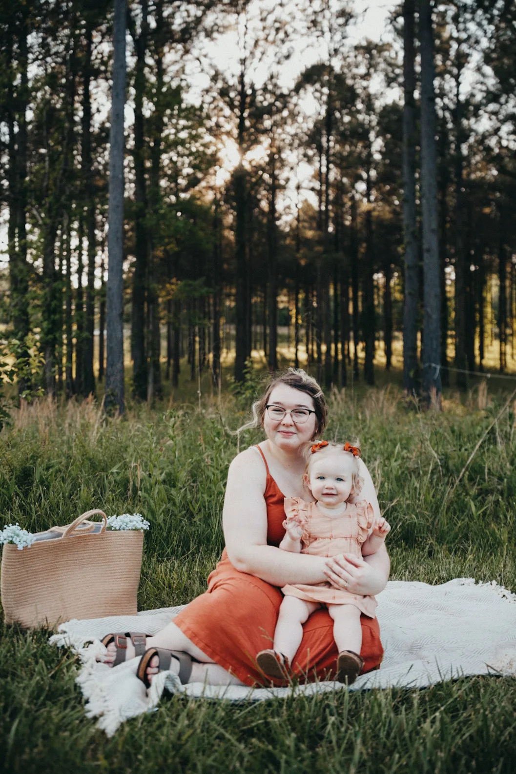 A woman dressed in an orange dress its on a white picnic balnket in a field her little girl in her arms