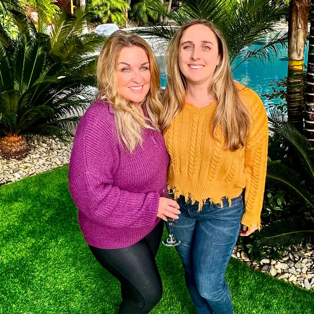 woman in pink top and black trousers and woman in orange top and jeans stood on grass outside.